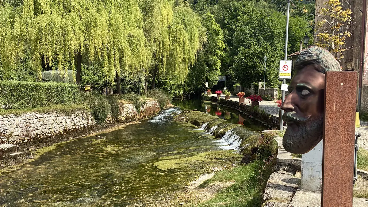Corso d’acqua alberato a Polcenigo con piccole cascate e scultura di volto in primo piano.