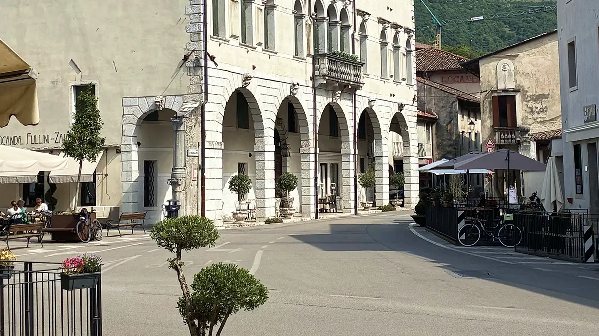 Piazza di Polcenigo con palazzo porticato, caffè all’aperto e biciclette.
